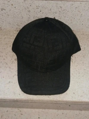 Svart keps från Fendi med mönster - Snygg svart keps från Fendi med diskret ton-i-ton FF-mönster över hela kepsen. Klassisk böjd skärm och justerbar baksida. Insidan har Fendi-loggor på banden. Perfekt accessoar för en trendig och exklusiv look.