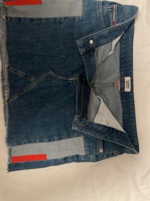 Jeanskjol från Tommy Hilfiger - Säljer en snygg jeanskjol från Tommy Hilfiger i klassisk blå denim. Kjolen har råa kanter, dekorativa sömmar och röda detaljer på sidorna. Den har fickor fram och bak samt knapp- och dragkedjestängning. Perfekt för en trendig och avslappnad look.