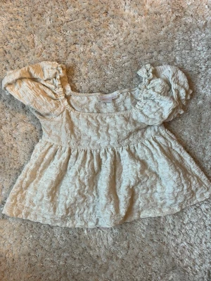 Vit blus med puffärm och struktur - Supersöt vit blus med puffärmar och volang nedtill. Toppen har en strukturerad yta med broderade detaljer och fyrkantig ringning. Perfekt för dig som gillar romantisk och trendig stil.