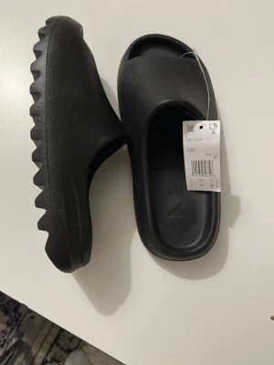 Svarta Yeezy Slide tofflor från Adidas - Säljer ett par svarta Yeezy Slide tofflor från Adidas. De har en chunky, räfflad sula och är tillverkade i ett mjukt, skönt syntetmaterial. Perfekta för en avslappnad och trendig look. Minimalistisk design med öppen tå och Adidas-logga på fotbädden.