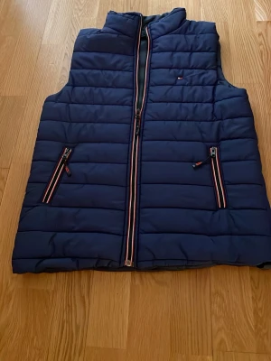 Marinblå dunväst Tommy Hilfiger - Snygg marinblå dunväst från Tommy Hilfiger med quiltad design och dragkedjor med röd, vit och blå detalj. Västen har hög krage och två fickor framtill. Perfekt för lager på lager och streetstyle.