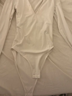 Vit långärmad body från H&M - Säljer en enkel och stilren vit body från H&M med långa ärmar och djup v-ringning. Tillverkad i mjukt stretchigt material som sitter snyggt på kroppen. Perfekt att matcha med jeans eller kjol för en clean look.