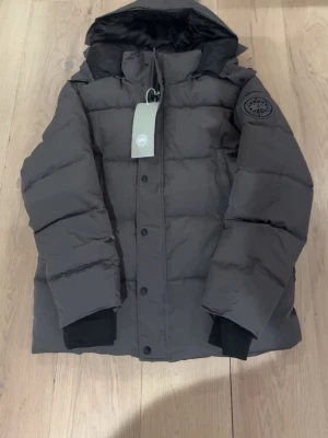 Grå pufferjacka från Canada Goose - Säljer en grå pufferjacka från Canada Goose med svart huva och emblem på ärmen. Jackan har breda kanaler, hög krage och svarta muddar vid ärmsluten. Perfekt för kalla dagar och riktigt snygg streetstil.