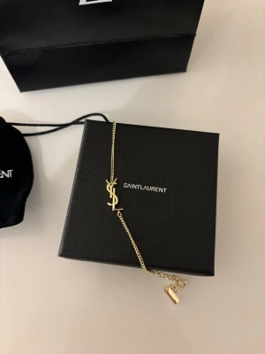 Guldigt halsband från Saint Laurent - Säljer ett elegant guldigt halsband från Saint Laurent med den ikoniska YSL-logotypen som hänge. Kedjan är tunn och stilren, perfekt för att lyfta vilken outfit som helst. Kommer med original ask och påse från Yves Saint Laurent.