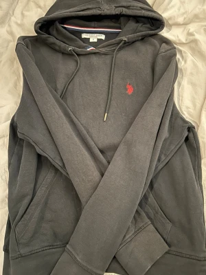 Mörkgrå hoodie från U.S.Pola Assn - Snygg mörkgrå hoodie från U.S.Polo Assn med klassisk röd broderad logga på bröstet. Hoodien har dragsko i huvan, känguruficka och ribbade muddar. Tillverkad i mjuk bomullsblandning för extra komfort.