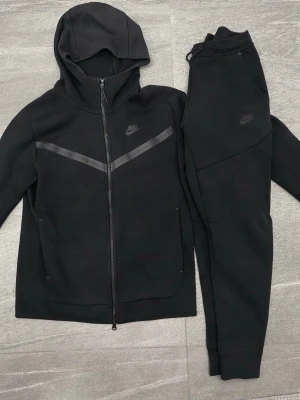 Nike tech fleece  - Svart Nike tech fleece dress med en huvtröja och matchande träningsbyxor. dressen är i sparsamt skick men behöver däremot en tvätt och då bör den se ny ut igen! Storlek S på tröja och XS på byxor