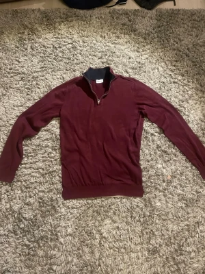 Vinröd halvzip  tröja - Säljer en vinröd stickad tröja med halv dragkedja och ståkrage i kontrasterande mörk färg. Tröjan har långa ärmar, ribbade muddar och är i ett mjukt, tunt material. Perfekt för lager-på-lager och snygg till jeans.