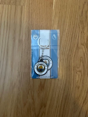 Manchester City nyckelring - Säljer en officiell Manchester City nyckelring i metall med klubbens klassiska logga i färgerna blå, vit, gul och röd. Perfekt accessoar för dig som vill visa ditt stöd för laget och ha en snygg detalj på nyckelknippan.