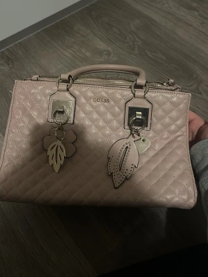 Ljusrosa quiltad handväska Guess - Säljer en ljusrosa quiltad handväska från Guess med silverfärgade metalldetaljer och dekorativa hängen i form av blad och blomma. Väskan har både handtag och avtagbar axelrem, samt dragkedja upptill. Perfekt för dig som gillar stilrena accessoarer.obbs finns en liten blå fläk från en penna men syns knappt!