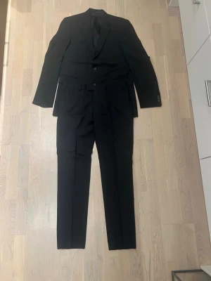 Svart kostym med tvåknäppt kavaj - Klassisk svart kostym med tvåknäppt kavaj och matchande byxor. Kavajen har smal passform, två framfickor med lock och en bröstficka. Byxorna har raka ben och bälteshällor. Perfekt för en stilren look.