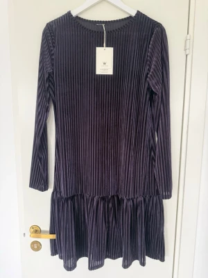 Mörklila ribbad klänning från F.A.V - Supersnygg mörklila klänning från F.A.V i ribbad sammet med lång ärm och volang nertill. Klänningen har rund halsringning och en kortare modell som ger en trendig look. Perfekt för dig som gillar att sticka ut med stilrena detaljer.