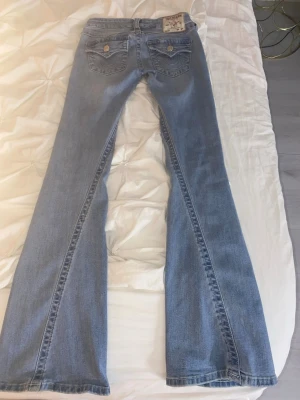 Ljusblå bootcut jeans från True Religion - Säljer ett par ljusblå bootcut jeans från True Religion med klassiska kontrastsömmar och ikoniska fickdetaljer bak. Jeansen har normal passform upptill och vida ben. Perfekta för dig som gillar retrovibbar och vill sticka ut lite extra.