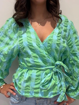 Grönblå omlottblus med knytband - Snygg omlottblus i grönt och blått rutmönster med knytband i midjan. Blusen har lång ärm och v-ringad hals. Materialet är lätt och krinklat, vilket ger en avslappnad look. Perfekt att styla med jeans för en trendig outfit.
