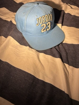 Ljusblå Jordan 23 keps - Snygg ljusblå keps från Jordan med broderad text 'JORDAN 23' framtill och en patch med Jumpman-loggan och siffran 23 på sidan. Klassisk böjd skärm och justerbar baksida. Perfekt för dig som gillar streetwear och sportig stil. Bara att fråga för fler bilder 