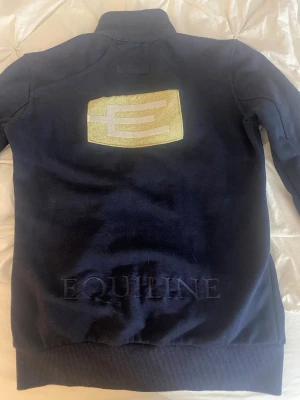 Marinblå ziptröja från Equiline - Snygg marinblå ziptröja från Equiline med hög krage och dragkedja framtill. Tröjan har två fickor, ribbade muddar och ett guldfärgat Equiline-emblem på ryggen samt broderad logga på bröstet. Perfekt för dig som gillar stilren design och hästsport.