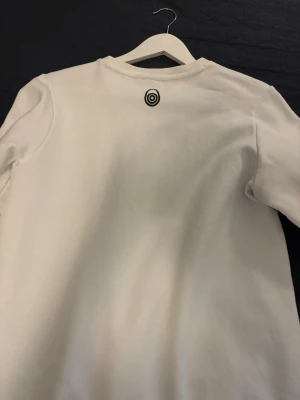 Vit sweatshirt från Sail Racing - Vit sweatshirt från Sail Racing med svart logotyp tryckt framtill och en liten logga i nacken. Tröjan har rund halsringning och är tillverkad i mjukt bomullsmaterial.