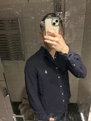 Mörkblå skjorta från Ralph Lauren - Snygg mörkblå skjorta från Ralph Lauren med klassisk krage och knappar framtill. Skjortan har lång ärm och den ikoniska broderade loggan på bröstet. Perfekt för en stilren look. Villig att gå ner lite i pris för snabb affär!