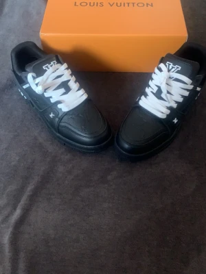 Louis Vuitton svarta sneakers - Louis Vuitton sneakers i svart läder med vita detaljer och vita skosnören. Klassisk LV-logga på plösen och sidan, samt ikoniska monogramdetaljer på sulan. Snygg och modern design med låg profil som passar till streetwear och trendiga outfits.