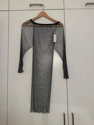 Svart nätklänning från Giorgia - Unik svart lång nätklänning från Giorgia med lång ärm och transparent design. Klänningen har ett grovmaskigt nätmönster och är perfekt för att skapa en edgy och trendig look. Bär den över andra plagg för en cool layering-effekt.