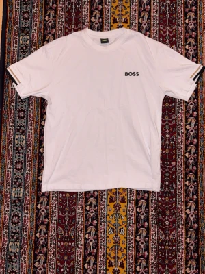 Hugo Boss T-shirt - Vit t-shirt från BOSS med svart broderad logga på bröstet och en snygg randig detalj längs ryggen. Ärmarna har också randiga detaljer. Klassisk passform och rund halsringning. Perfekt för en clean och stilren look. Är M men passar också S