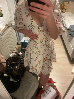 Blommig kortklänning med v-ringning - Söt kortklänning i ljus färg med blommigt mönster och v-ringad hals. Klänningen har puffiga korta ärmar och markerad midja med en knytning på ryggen. Perfekt för varma dagar och somriga tillfällen. Från hm och i strolek XS, aldrig använd med lappen kvar. 