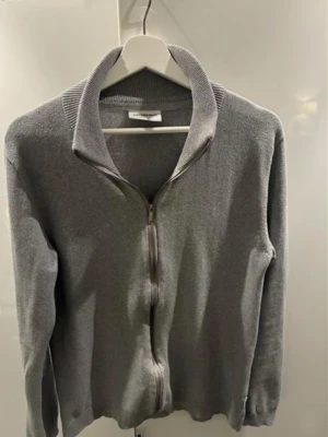 Grå ziptröja från j Lindbergh - Stilren grå ziptröja från j lindbergh  med hög krage och hel dragkedja framtill. Tröjan har ribbade muddar vid ärmslut och nederkant, samt en mjuk och tunn stickning som gör den enkel att matcha till olika outfits.