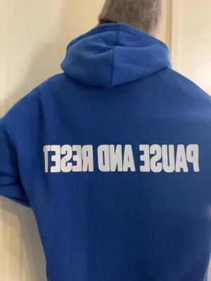 Blå hoodie med text  - Blå hoodie med stor vit text 'PAUSE AND RESET' på ryggen. Klassisk känguruficka framtill och huva. Tillverkad i mjukt material, perfekt för chill dagar. Ingen synlig logga eller märke.