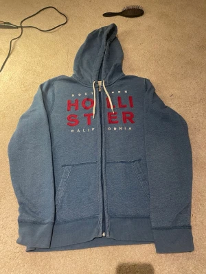 Blå zip hoodie från Hollister - Blå hoodie från Hollister med dragkedja och stor huva. Framtill står det 'Hollister California' i rött och vitt. Hoodien har kängurufickor och vita dragsnören. Perfekt för chill dagar och streetwear-stil.