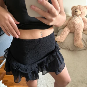 Svart volangkjol med resårmidja - Supersöt svart kortkjol med bred smockresår i midjan och dubbla volanglager i tunn, luftig polyester. Kjolen har en flowy känsla och passar perfekt till en trendig vardagslook.