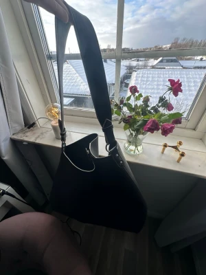 Svart axelväska i skinnimitation - Snygg svart axelväska med bred axelrem och silverfärgade metalldetaljer. Väskan har en mjuk, slouchy form och är tillverkad i skinnimitation. Perfekt för dig som vill ha en stilren och enkel väska med plats för det viktigaste.