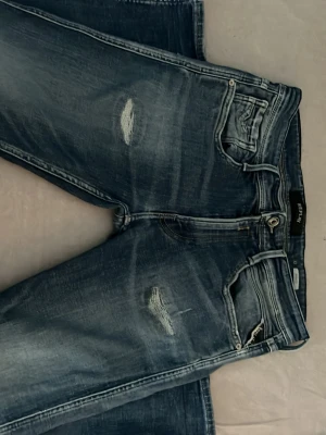 Blå slitna jeans från Replay anbass - Säljer ett par blå jeans från Replay i modellen Anbass. Jeansen har en snygg tvätt med slitningar på låret och vid fickan. Klassisk femficksmodell med dragkedja och knapp. Perfekta för en avslappnad look. Väldigt bra skick. W32 L30