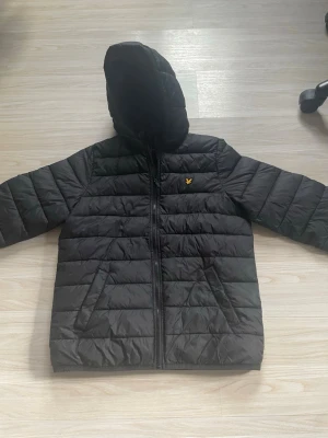 Svart dunjacka med huva Lyle & Scott - Säljer en svart tunn dunjacka från Lyle & Scott med huva och dragkedja framtill. Jackan har två snedställda fickor och det klassiska gula logomärket på bröstet. Quiltad design som ger en sportig och clean look, perfekt för kalla vår dagar