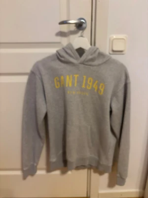 Grå hoodie från Gant med tryck - Snygg grå hoodie från Gant med gul text 'GANT 1949 NEW HAVEN' på bröstet. Klassisk modell med huva och magficka, ribbade muddar vid ärmslut och nederkant. Perfekt för en avslappnad och sportig stil.