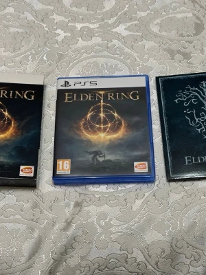 Elden Ring PS5 med fodral och extras - Elden Ring till PS5, komplett med blått plastfodral, originalskiva och extra material som artbook och specialomslag. Omslaget har en mörk design med gyllene symbol och fantasytema. Perfekt för dig som gillar action-RPG och episka äventyr.