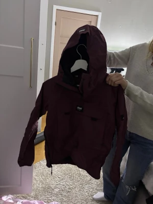 Vinröd vindjacka från Dope - Snygg vinröd vindjacka från Dope med huva och dragkedja. Jackan har en stor ficka framtill med Dope-logga, justerbara muddar och dragsko nertill. Perfekt för blåsiga dagar och outdoor-aktiviteter.