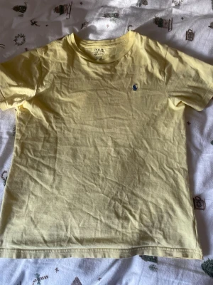 Gul t-shirt från Polo Ralph Lauren - Snygg gul t-shirt från Polo Ralph Lauren med klassisk blå logga broderad på bröstet. T-shirten har rund halsringning och korta ärmar, tillverkad i mjuk bomull som känns skön mot huden. Perfekt för en avslappnad och stilren look.