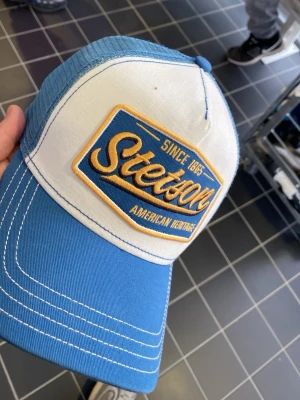 Blå och vit keps från Stetson - Snygg blå och vit keps från Stetson med broderad gul och blå logotyp framtill. Klassisk trucker-modell med mesh baktill och böjd skärm. Perfekt accessoar för en avslappnad streetstil.