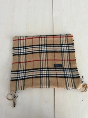 Beige rutig halsduk från Burberry - Klassisk beige halsduk från Burberry med ikoniskt rutmönster i svart, vitt och rött. Halsduken har fransar i ändarna och en vävd Burberry-etikett. Perfekt accessoar för att lyfta din outfit med en tidlös touch.