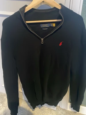 Svart tröja med dragkedja från Polo Ralph Lauren - Svart långärmad tröja från Polo Ralph Lauren med halv dragkedja vid halsen och röd broderad logga på bröstet. Tröjan har ribbade muddar och är tillverkad i ett stickat material som ger en klassisk och stilren look.