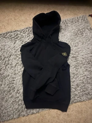 Svart hoodie från Stone Island - Svart hoodie från Stone Island med klassisk huva och dragsko. Ikonisk logopatch på vänster ärm. Mjuk bomullskvalitet och ribbade muddar vid ärmslut och nederkant. Perfekt för en stilren och avslappnad look.