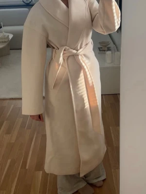 Beige lång kappa med bälte - Lång beige kappa från Ginatricot. Slutsåld nu. Köpt för 899kr och säljs för 599kr. Oanvänd. 