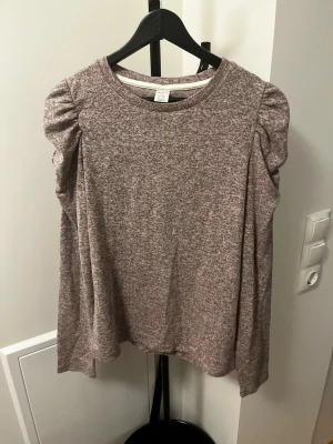 Beige/lila långärmad topp med puffaxel - Mysig beige/lila långärmad topp med lätt melerad look och puffade axlar. Rund halsringning och avslappnad passform. Perfekt för dig som gillar en enkel men trendig stil.