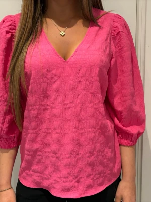 Rosa blus med v-ringning - Trendig rosa blus med v-ringad hals och trekvartsärmar. Blusen har en lätt strukturerad yta och puffiga ärmar med resår vid ärmslut. Perfekt för dig som vill ha en färgstark och bekväm topp till din outfit i sommar 