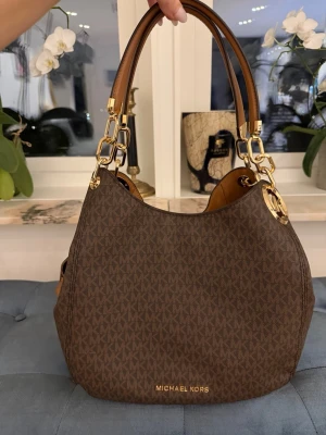 Brun Michael Kors handväska - Snygg brun handväska från Michael Kors med guldiga metalldetaljer och dubbla handtag i brunt läder. Väskan har MK-logga över hela ytan, dragkedjeficka på sidan och en rundad form. Perfekt för dig som gillar klassisk design och exklusiva detaljer.