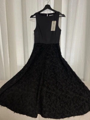 Svart långklänning med strukturkjol - Elegant svart långklänning med ärmlös överdel och vid kjol i strukturerat tyg. Klänningen har rund halsringning och knapp i nacken. Kjolen har en unik textur som ger en lyxig känsla. Perfekt för dig som vill sticka ut med stil.