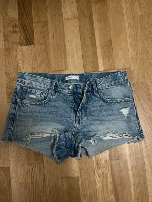 Blå slitna jeansshorts Perfect Jeans - Säljer ett par blå jeansshorts från Perfect Jeans (gina tricot) i storlek 36. Shortsen har slitna detaljer och fransar vid bensluten för en avslappnad look. Klassisk femficksmodell med knapp och dragkedja framtill. Perfekta till sommaren!