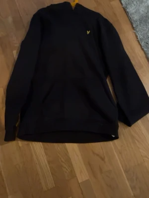 Svart hoodie från Lyle & Scott  - Säljer en svart hoodie från Lyle & Scott Junior med klassisk gul logga på bröstet. Hoodien har huva och känguruficka framtill. Tillverkad i mjukt material som passar perfekt för en avslappnad stil.
