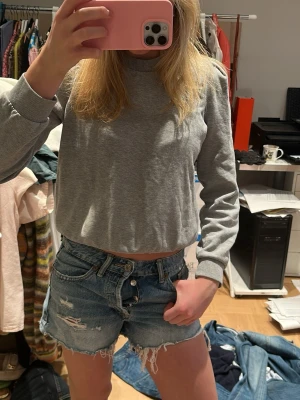 Blå slitna jeansshorts med fransar - Säljer ett par blå jeansshorts med råa, fransiga kanter och slitningar framtill. Klassisk femficksmodell med knappgylf och hög midja. Perfekta för en avslappnad och trendig look. Säljer pga att dem är för små