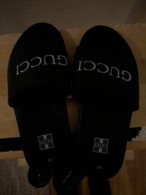 Svarta tofflor - Säljer ett par svarta tofflor med tydlig logga över foten. Klassisk slip-on modell med bred rem och räfflad sula för bra grepp. Perfekta för chill dagar eller när du vill ha en clean och lyxig look. Akopia därav priset