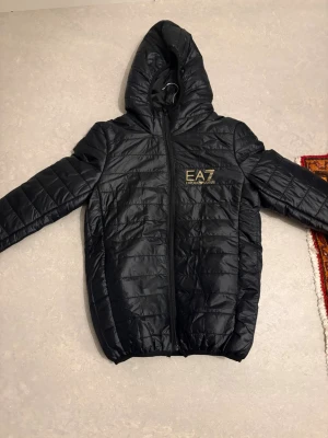 Svart pufferjacka från EA7 Emporio Armani - Säljer en svart pufferjacka från EA7 Emporio Armani med huva och guldfärgad logga på bröstet. Jackan har quiltad design, dragkedja framtill och långa ärmar. Perfekt för dig som vill ha en lätt och snygg jacka till höst och vinter. Inte äkta